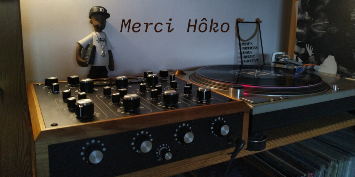 Table de mixage (Carmen SE) et platine vinyle, avec un disque, et à l'arrière une figurine de J Dilla, le tout faiblement éclairé. Il est écrit « Merci Hôko » en surimpression.